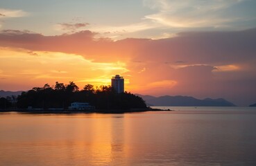 Island silhouette at sunset with vibrant orange sky and calm sea reflections. A tall building stands on the tree covered island shore. Distant mountains fade into twilight mist.