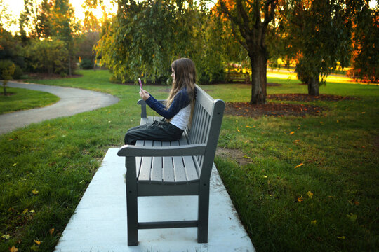 Profile young girl looking at cell phone on park bench sunny day