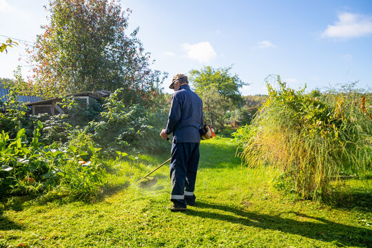 Senior man uses grass trimmer to cut overgrown lawn in his backyard