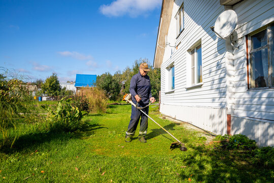 Senior man trimming overgrown grass with string trimmer around house