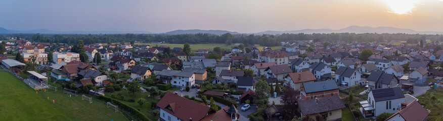 Fototapeta premium Panoramic aerial view of Ihan, Slovenia at sunset, showing a peaceful residential village with family houses, green fields, football pitch, trees and distant hills under warm evening light.