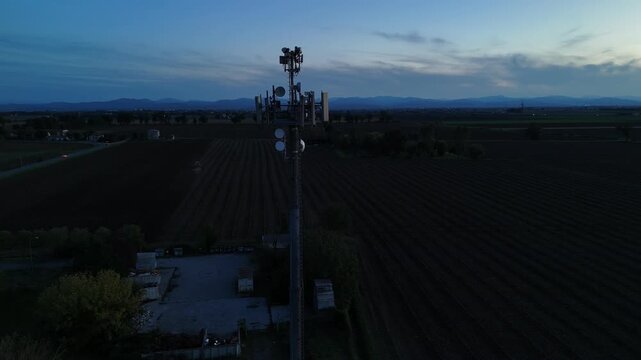 Aerial footage of a tall telecommunication tower equipped with antennas and satellite dishes, transmitting signals against a clear dark blue sky at dusk in a rural landscape