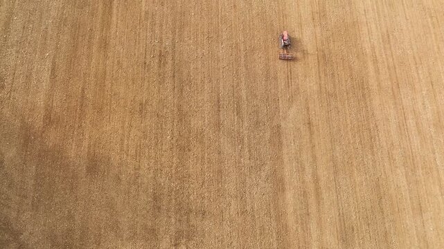 Bird's eye view of a tractor equipped with a harrow system tilling the soil in a large, arid agricultural field on a bright spring day, getting ready for planting crops