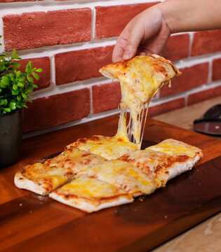 A slice hot traditional pinsa romana pizza square shaped toppings with stretching cheese golden yellow color on wooden cutting board and brick wall tasty snack fresh delicious gourmet italian cuisine.
