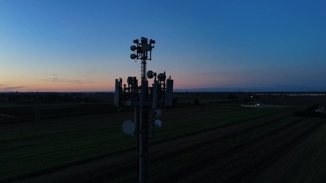 Aerial footage of a tall telecommunication tower equipped with antennas and satellite dishes, transmitting signals against a clear dark blue sky at dusk in a rural landscape