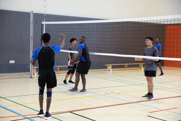 Obraz premium Diverse male teenage athletes playing volleyball in gym, holding ball near net, wearing knee pads