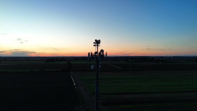Aerial footage of a tall telecommunication tower equipped with antennas and satellite dishes, transmitting signals against a clear dark blue sky at dusk in a rural landscape