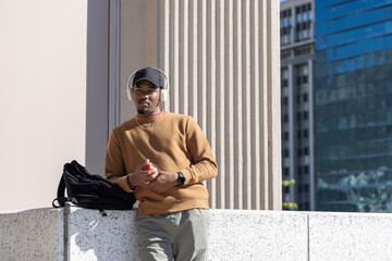 African American man leaning on ledge in plaza, wearing headphones, holding red phone, copy space © wavebreak3