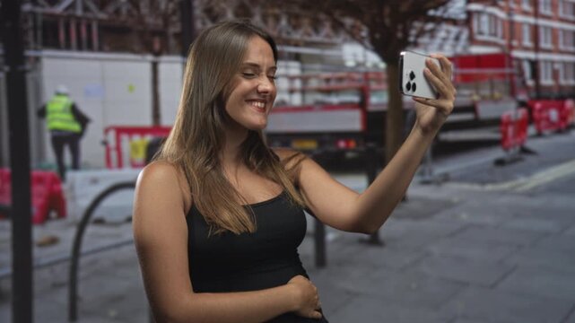 Woman holding smartphone for selfie on a street, arm raised and smiling, wearing a black tank top and standing near construction barriers; happy candid moment.