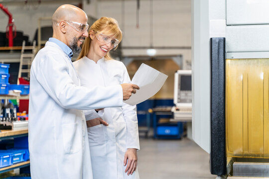 Two happy technicians wearing lab coats and safety glasses looking at plan in factory