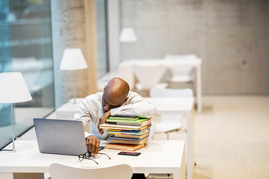 Exhausted mature man sitting at desk leaning on stack of books