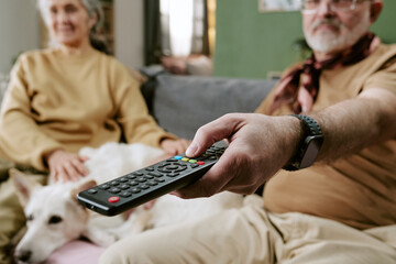 Senior Caucasian man holding television remote control while sitting on sofa next to senior...