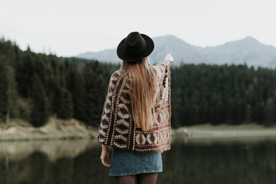 Back view of fashionable young woman wearing hat and poncho standing in front of a lake
