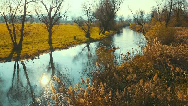 Kayakers navigate a serene, sunlit river surrounded by golden reeds and bare trees, capturing the essence of peaceful outdoor adventure. The long shot, tranquil setting evokes a sense of escape.