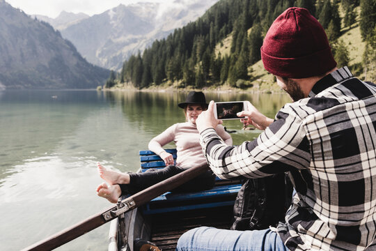 Austria, Tyrol, Alps, man taking cell phone picture of woman in rowing boat on mountain lake