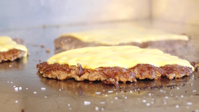 Close-up of beef burger patties cooking on a hot griddle, topped with melting cheese slices. Steam rises from the surface, highlighting texture and juicy detail. 