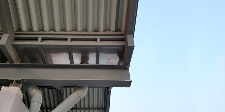 Modern industrial roof structure featuring gray corrugated metal sheets sturdy steel beams and professional drainage system with rain gutters and PVC pipes against clear blue sky