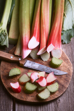 Rhubarb chunks scattered on wooden board with kitchen knife, natural light food styling. Organic fresh rhubarb slices detail, healthy vegetable ingredient