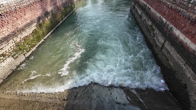 Wide-angle view of a rural irrigation canal with a small brick masonry check dam and rushing white water in a forested area of Haryana, India.