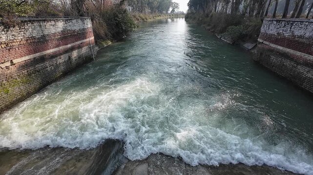 Wide-angle view of a rural irrigation canal with a small brick masonry check dam and rushing white water in a forested area of Haryana, India.