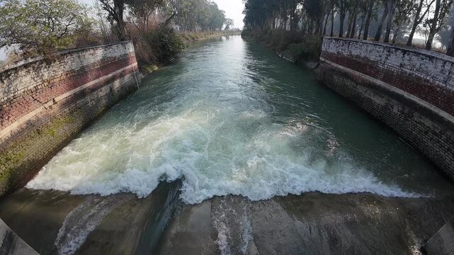 Wide-angle view of a rural irrigation canal with a small brick masonry check dam and rushing white water in a forested area of Haryana, India.