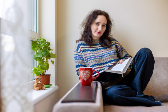 Middle-aged woman relaxing with book and mug. Cozy lifestyle portrait of a middle-aged brunette woman sitting on a sofa by the window, reading a book with a red mug nearby.