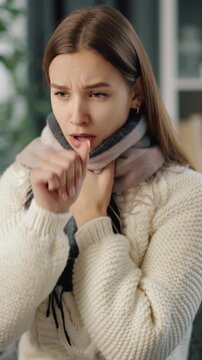 Woman wearing a scarf coughs while sitting in a cozy home.