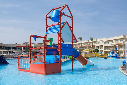 Colorful water play structure with slides and fountains in a resort pool area, surrounded by palm trees and hotel buildings
