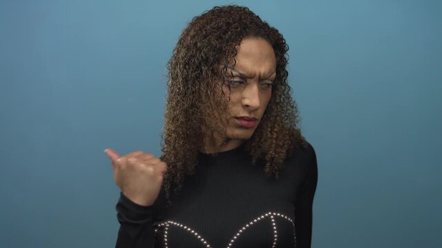 Transgender woman pointing her thumb sideways against a solid blue studio backdrop with a furrowed brow; confusion.
