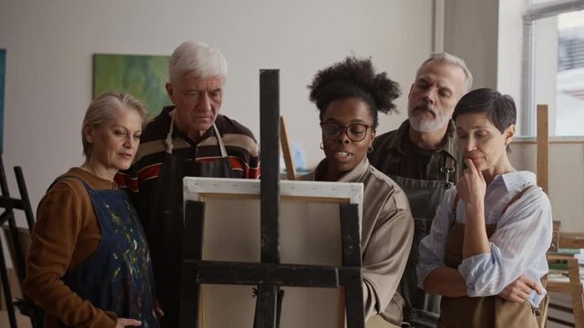 Group of senior students standing with Black female artist near easel painting and talking about art in bright studio