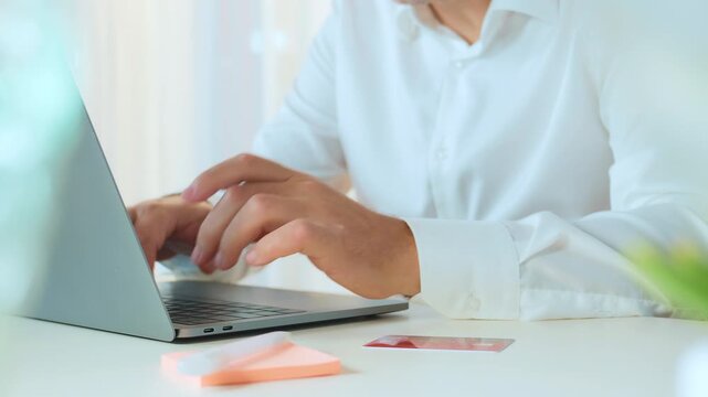 A person in a white shirt sits at a desk, holding a credit card and using a laptop. Nearby are more cards and cash, indicating online shopping or financial transactions.