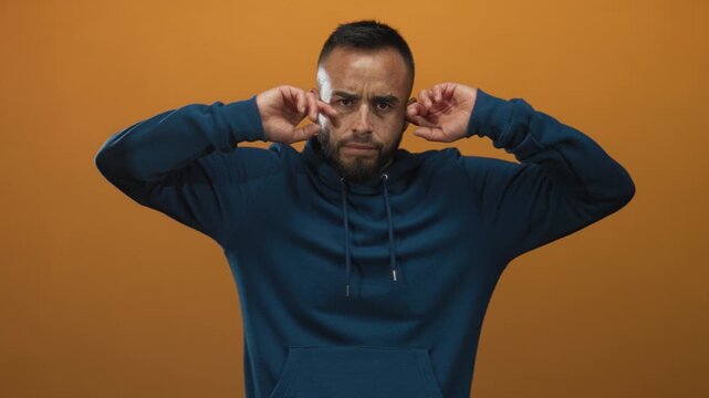 Young hispanic man wearing blue drawstring hoodie and gold earring covering ears with fingers in orange studio; frustration.