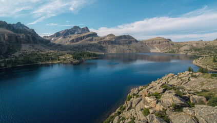 Obraz premium Lucid deep blue mountain lake with rocky shore and distant peaks under blue sky