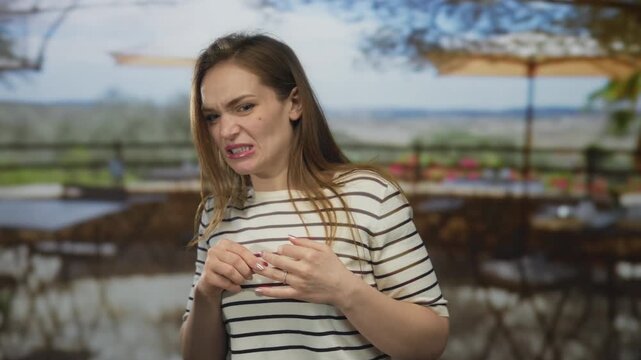 Woman in striped shirt grimaces with clenched fists and scrunched nose on sunlit restaurant terrace outdoors; disgust.