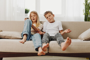 Two barefoot children sitting on a beige sofa in a bright living room, relaxed casual clothing. Concept of childhood, family, and home lifestyle with natural light and cozy atmosphere. © SHOTPRIME STUDIO