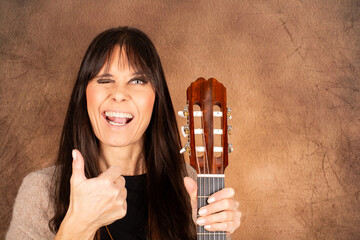 Fr&ouml;hliche Frau mit Akustikgitarre zwinkert und zeigt Daumen hoch vor warmem Hintergrund&ldquo;