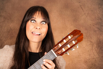 Humorvolle Frau mit Akustikgitarre macht lustigen Gesichtsausdruck vor braunem Studiohintergrund
