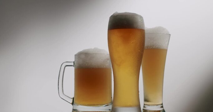 Different types of beer glasses, including a pint, a mug, and a pilsner glass, standing with foamy heads on a white background with a subtle shadow. Three different beer glasses with foam