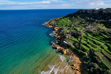 Fototapeta premium Aerial drone view of the most beautiful Malta beach in Gozo, vibrant red sand Ramla beach with crystal clear blue water and mesmerizing waves with lush green vegetation around it and the famous Mixta 