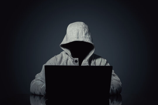 Faceless hooded figure using laptop on dark background, anonymous hacker concept, cybercrime, data breach, online security threat, internet blocking, privacy risk, digital attack scene at night