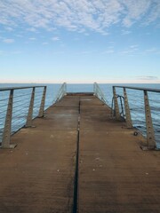 Fototapeta premium A long, narrow wooden pier stretches out toward a calm sea under a clear blue sky with soft, wispy clouds. The symmetrical perspective creates a sense of peaceful solitude and infinity.