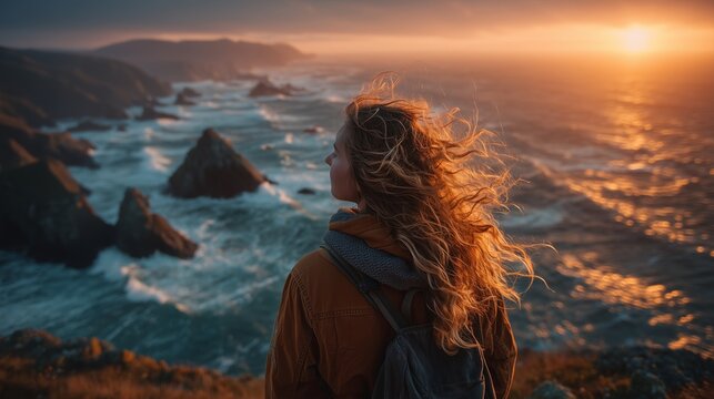 Female traveler standing on a cliff overlooking a vast ocean, wind flowing through her hair at sunset.