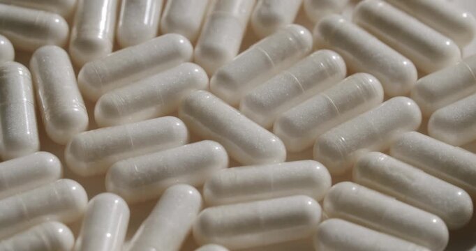 Extreme close-up of a large number of white translucent pill capsules with powder inside slowly piling up, suggesting pharmaceutical manufacturing or nutritional supplement production concepts