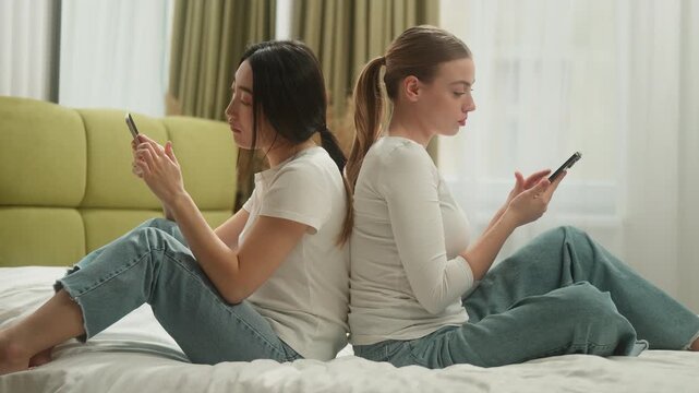 Two diversity women on bed leaning backs chatting mobile phones after conflict offended girls friends Caucasian Asian Korean Japanese Chinese female addicted internet browsing smartphones at home