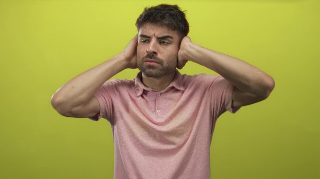 Young hispanic man covers ears with both hands in vibrant green studio set while grimacing in discomfort; annoyance.