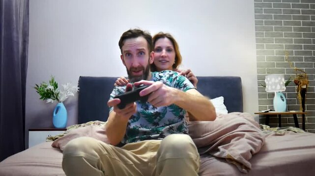 A young man with a joystick plays a console while sitting on a double bed and a sexy girl harasses him from behind