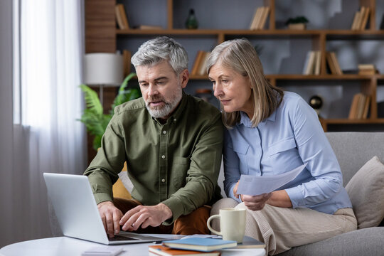 Mature couple on sofa in cozy living room using laptop and papers to review bills, budget and retirement plans, focused on savings, mortgage and investments for future