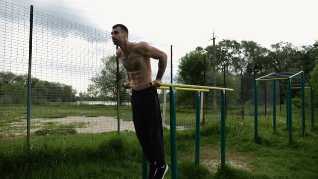 Young sportsman doing push ups on parallel bars at nature. Strong sporty man exercising at sports ground. Male shirtless athlete training hard outdoor. Concept of active sportive lifestyle. Slow mo