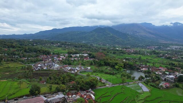 Aerial drone footage of a green valley, with many villages, surrounded by forests, agricultural fields, rice fields, volcano mountains, in cloudy atmosphere, in Garut area, Java island, Indonesia