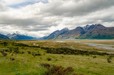 Fototapeta premium Rangitata River, with Mount Cook and surrounding mountains in the background. The scene includes a lush green foreground with shrubs and rolling hills, along with cloudy skies.